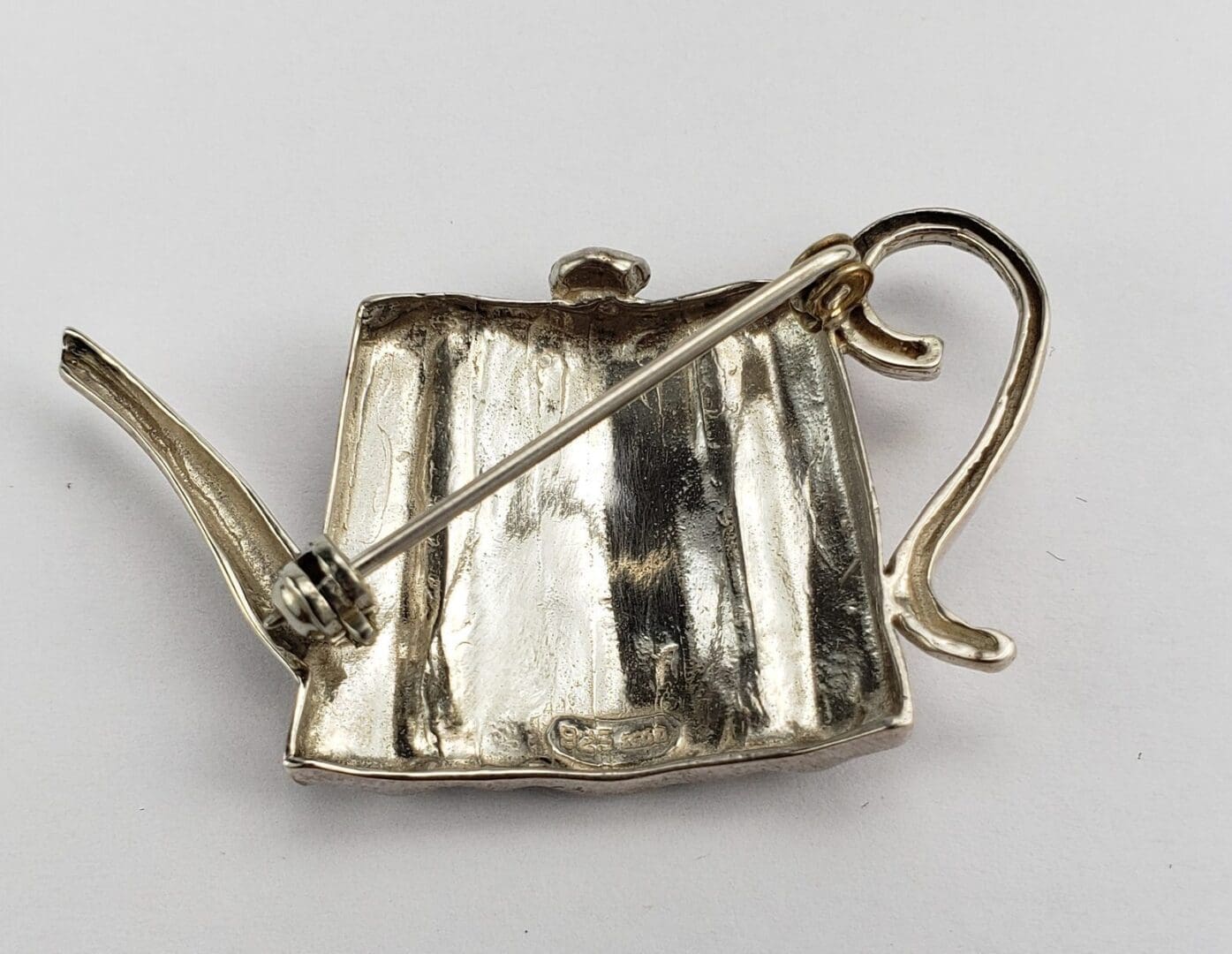 Silver teapot-shaped brooch on white background.