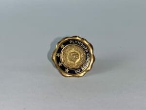 Gold Plymouth Corporation pin on gray background.